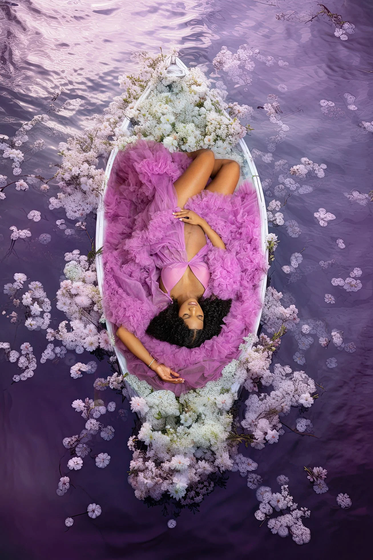 A woman lying in a small boat decorated with pink and white flowers is floating on purple water, surrounded by floating blossoms.