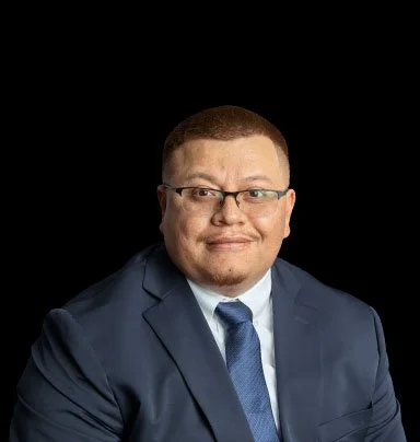 Portrait of a man in a navy suit, white shirt, and blue tie, wearing glasses, against a black background.
