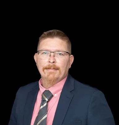 A man with glasses, a beard, and a mustache wearing a navy suit, pink shirt, and striped tie against a black background.