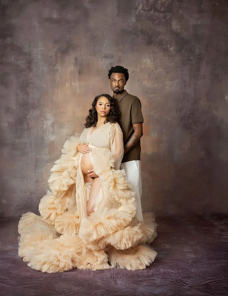 A pregnant woman in a flowing, ruffled beige gown gently holding her belly, standing beside a man in a brown polo shirt and white pants against a textured backdrop.