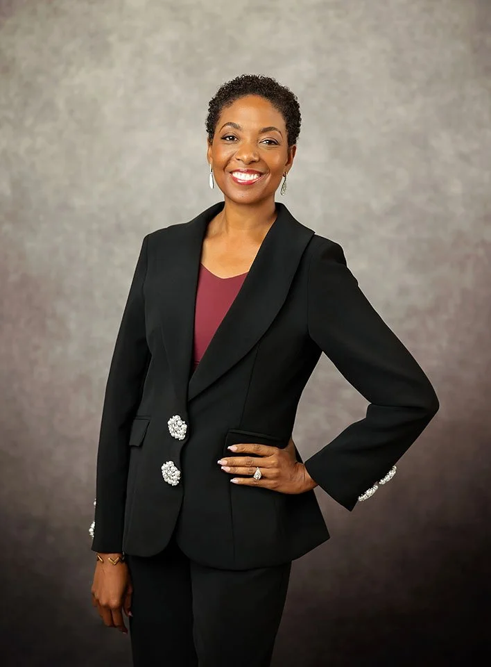 A woman in a black blazer with decorative buttons, wearing a maroon top, smiling and posing with her hand on her hip against a gradient gray background.