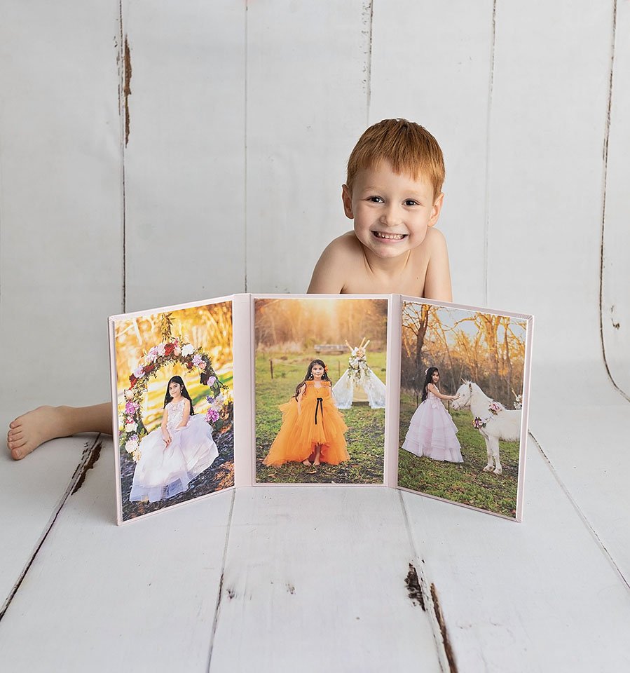 A young boy with red hair smiling and sitting behind a tri-fold photo display of three images of girls in colorful dresses outdoors.