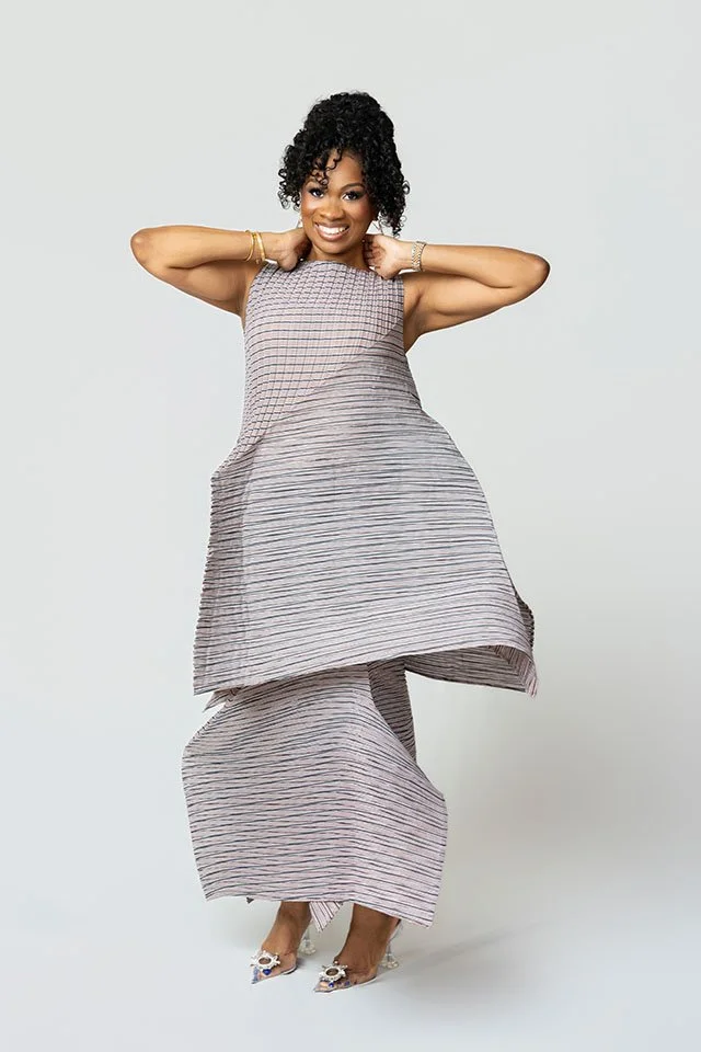 A woman with curly black hair smiling, wearing a layered, striped dress and high heels, standing against a plain white background.