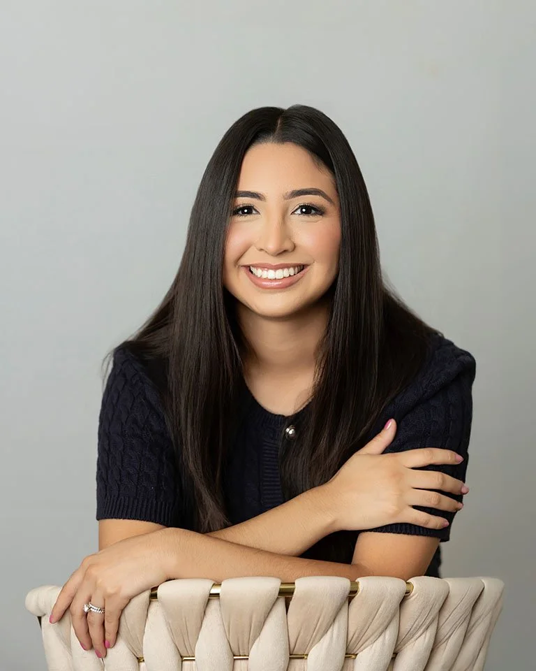 A smiling woman with long dark hair, wearing a dark short-sleeve sweater, sitting on a cream-colored chair with a light gray background.