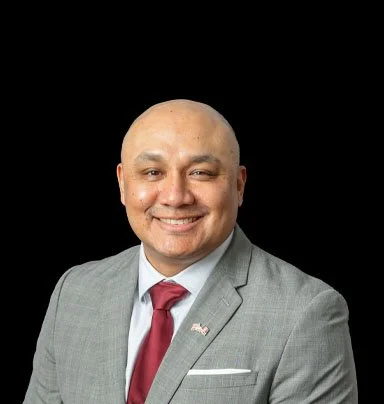Portrait of a bald man in a gray suit with a red tie and American flag pin, smiling against a black background.