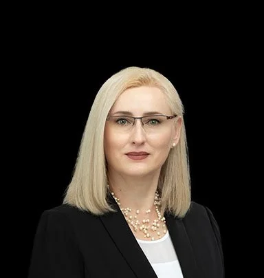 Professional woman with shoulder-length blonde hair, wearing glasses, a black blazer, and a pearl necklace, posing against a black background.