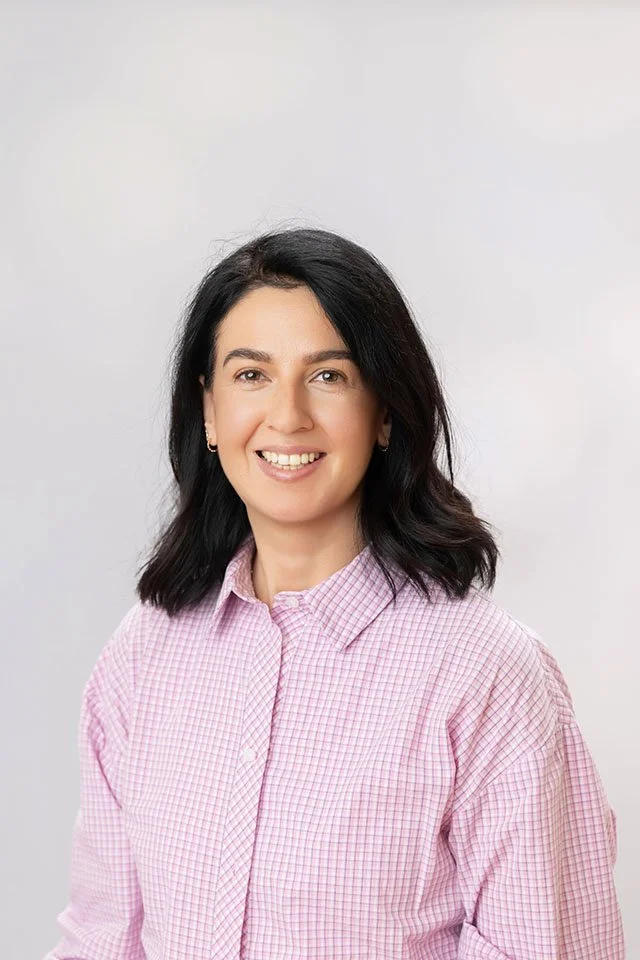 A woman with dark hair and light skin, smiling, wearing a pink checkered shirt, against a plain light gray background.