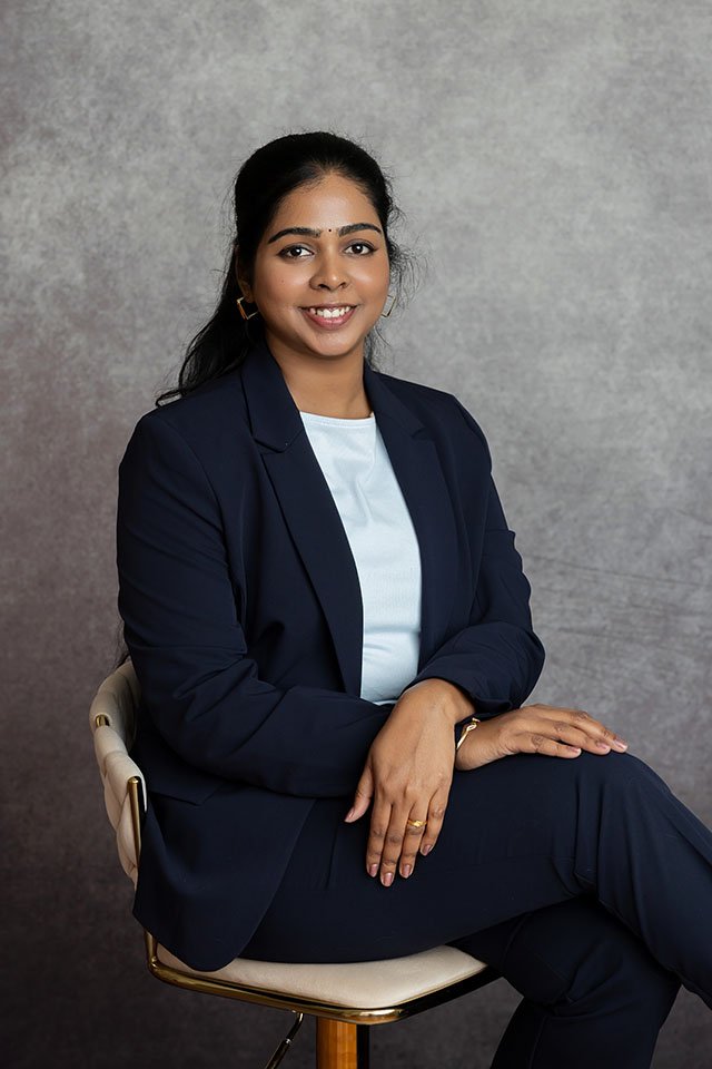A woman sitting on a chair, wearing a navy blue blazer over a white top, smiling, with a gray background.