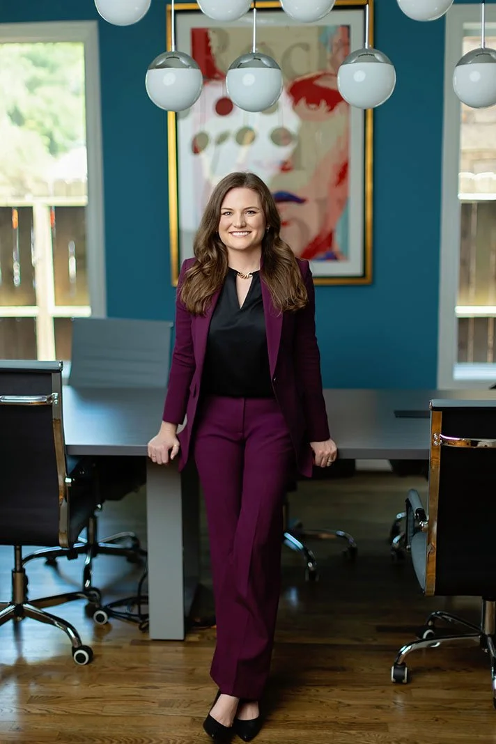 A woman in a purple suit standing in a modern conference room with a large colorful portrait of a face on the wall behind her.