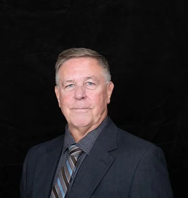 A middle-aged man with gray hair wearing a dark suit, patterned shirt, and striped tie, standing against a black background.