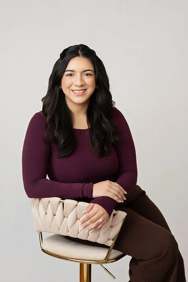 A woman with long dark hair smiling and sitting on a beige upholstered chair with a woven design, dressed in a maroon long-sleeve shirt and brown pants, against a plain light gray background.