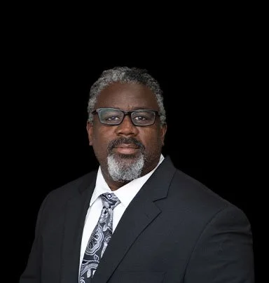 A middle-aged African American man with gray hair and beard, wearing glasses, a black suit, white shirt, and patterned tie, against a black background.