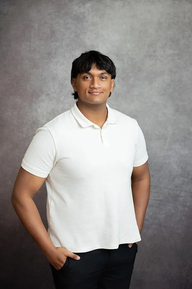 A man with black hair and medium skin tone wearing a white polo shirt and black pants, standing with hands in pockets against a gray textured background.