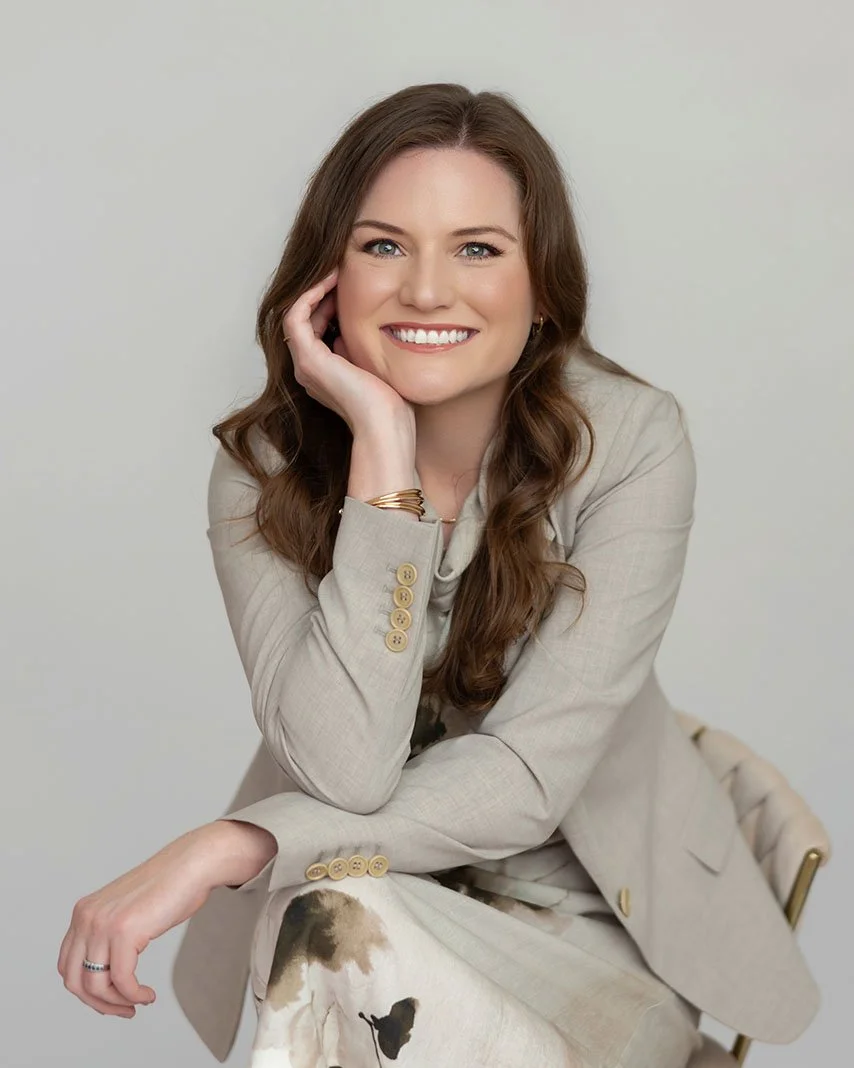 A woman with long brown hair, smiling and resting her chin on her hand, wearing a beige blazer and patterned pants, sitting against a plain light background.