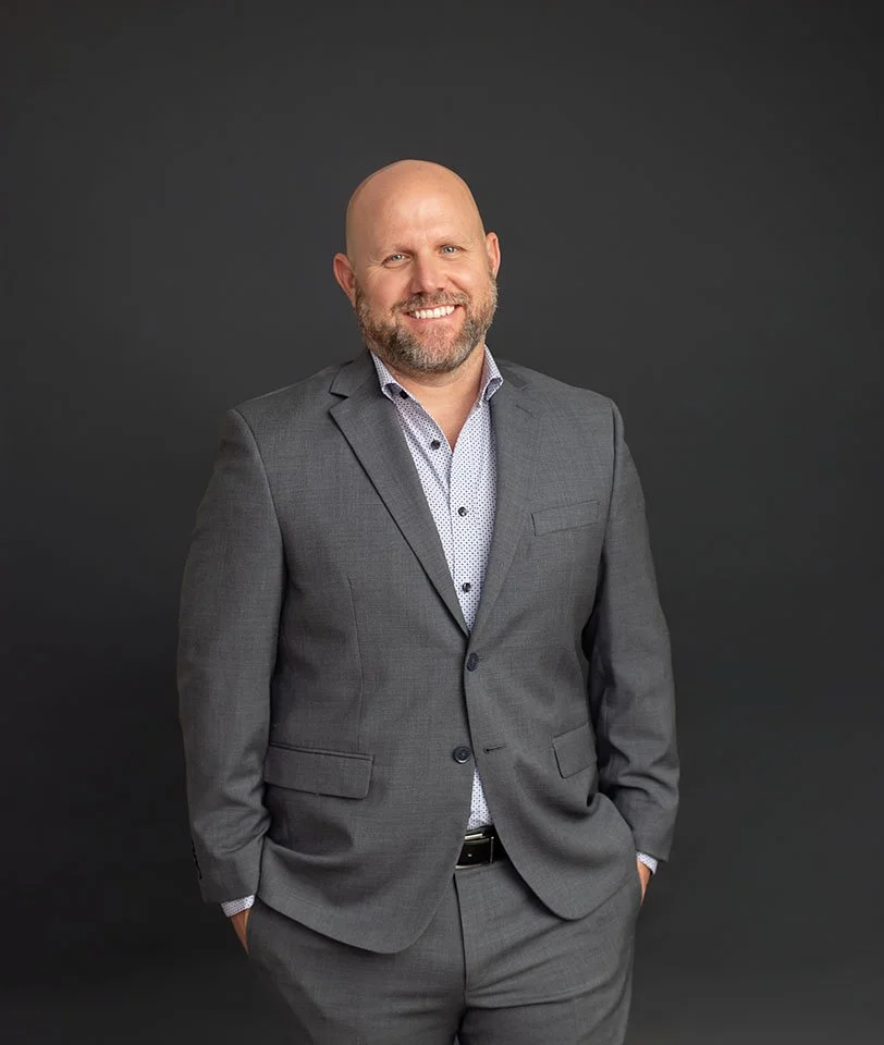 A bald man with a beard wearing a gray suit and a patterned shirt, smiling and standing against a dark gray background.