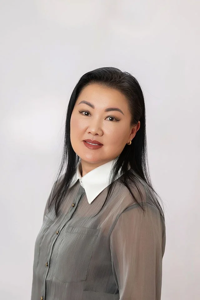 Portrait of an Asian woman with long dark hair, wearing a sheer gray blouse with gold buttons and a white collar, smiling softly against a plain white background.