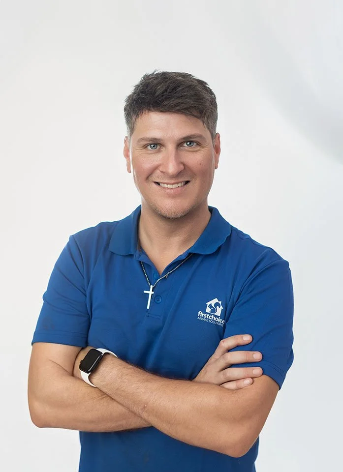 A smiling man in a blue polo shirt with a logo, wearing a cross necklace and a smartwatch, standing with arms crossed against a plain white background.