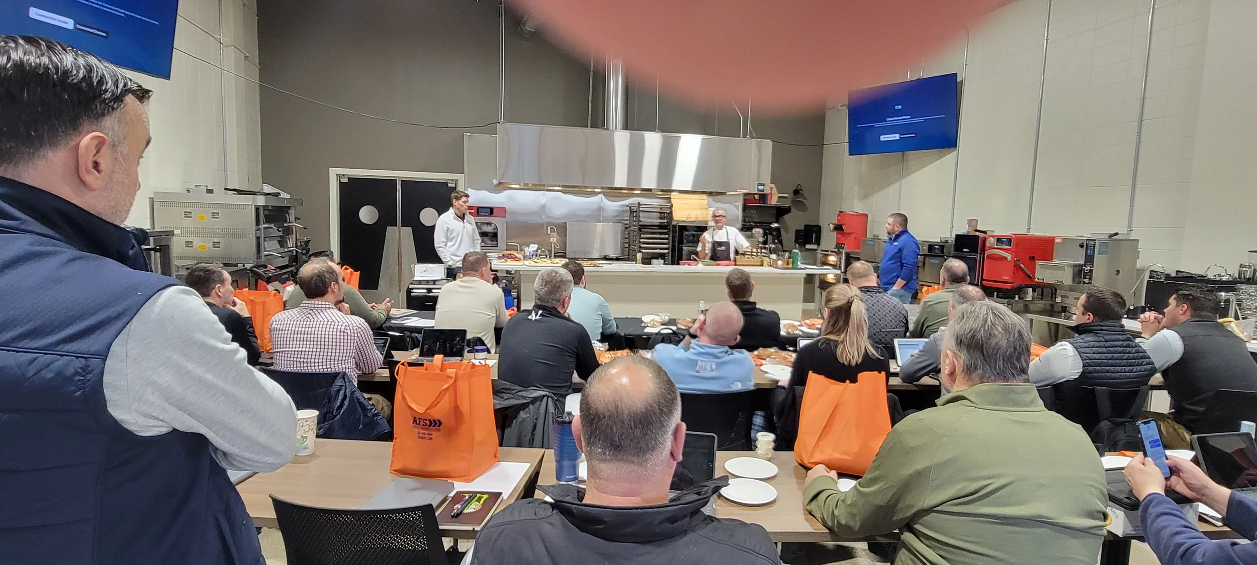 Large group attending a culinary equipment demonstration in a commercial kitchen classroom with chef instructors presenting at the front.