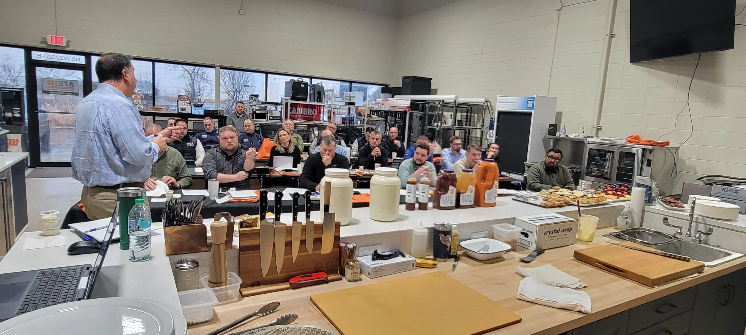 Instructor leading a foodservice training class in a commercial test kitchen with attendees seated at tables and cooking tools on the counter.