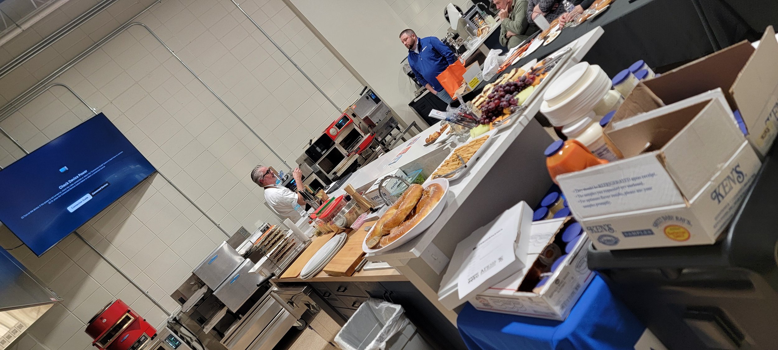 Chef demonstrating food preparation in a commercial kitchen training room with ovens, prep tables, and food samples displayed on the counter.