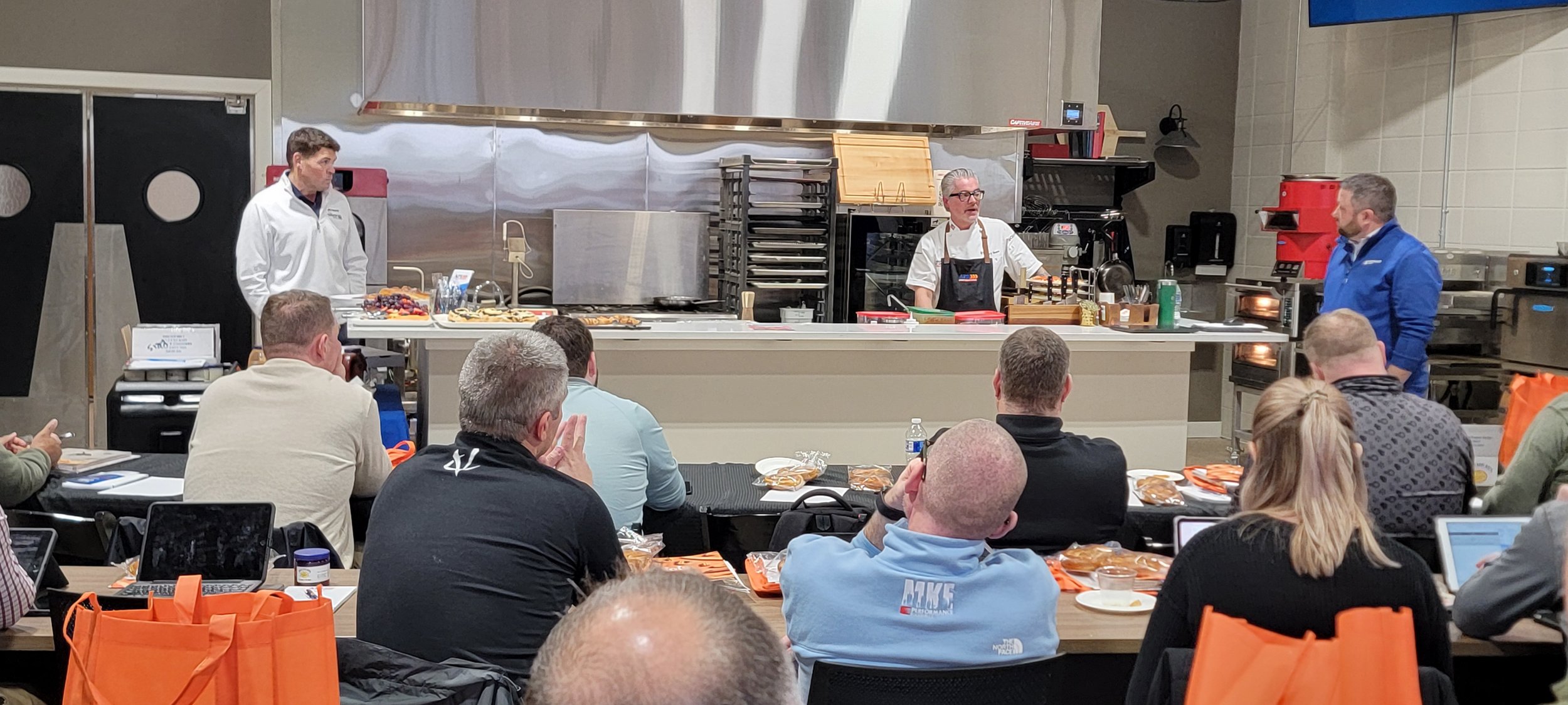 Chef presenting cooking techniques to a group of foodservice professionals during a kitchen equipment demonstration with prepared food on display.
