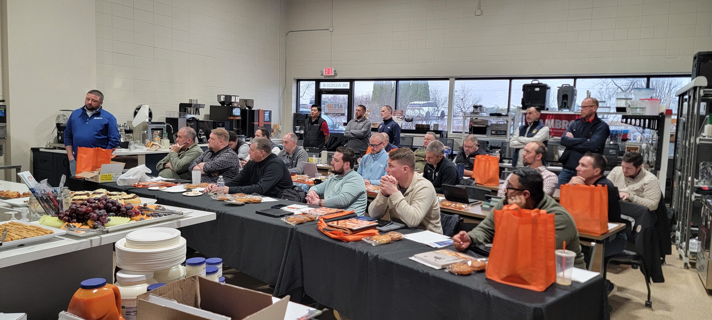 Group of foodservice professionals attending a training session in a kitchen showroom with food samples, equipment, and orange tote bags on tables.