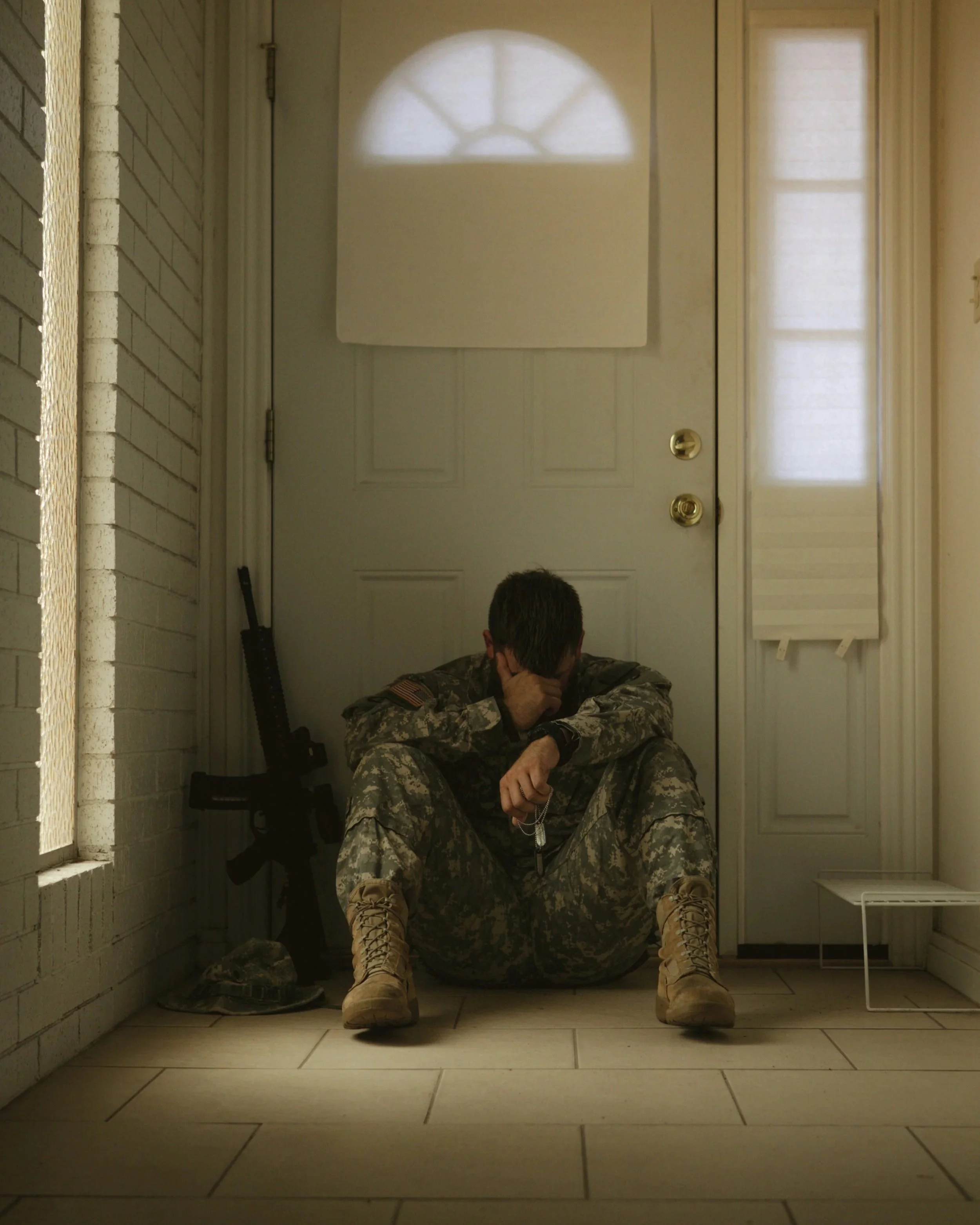 Solider sitting on the floor leaning against the front door of his house with his head in his hands , rifle leaning against the wall
