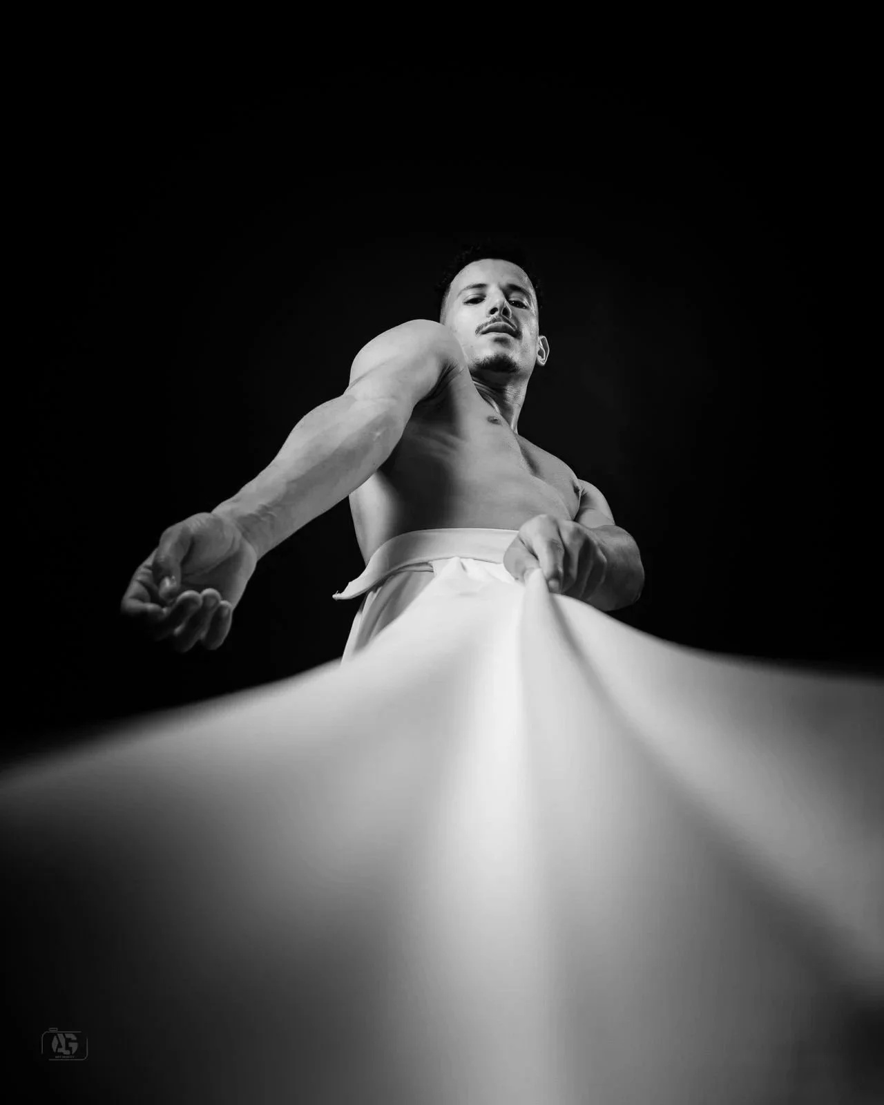 Low-angle shot of a shirtless man pulling on a piece of white fabric, against a dark background. Karim El Enoury