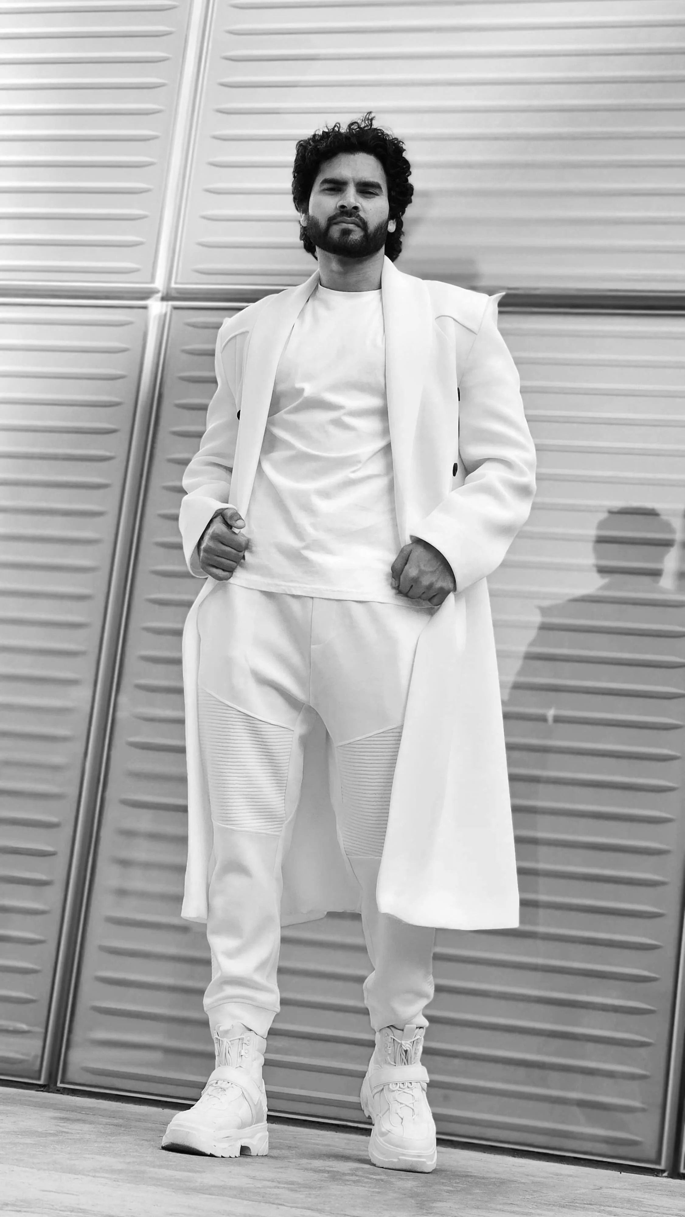 Man in an all-white outfit with a long coat, standing against a modern metallic wall. Jeetu Singh