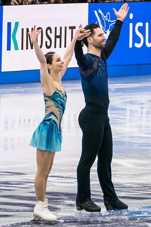 Deanna Stellato-Dudek and Maxime Deschamps, two figure skaters with their arms raised at the end of the pairs free skate at the 2025 World Figure Skating Championships.