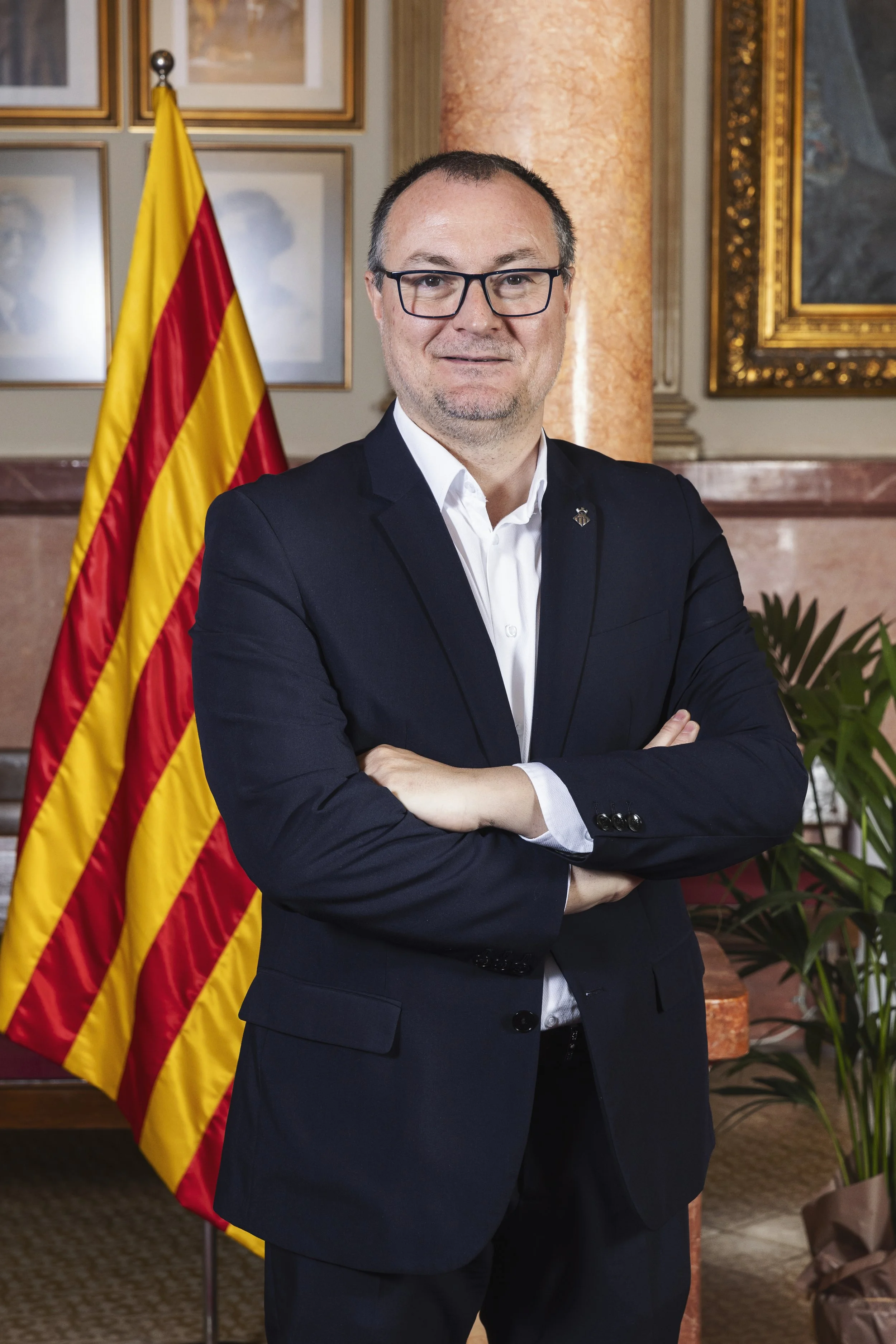 Hombre con gafas de vestir con brazos cruzados frente a la bandera de España en un recinto con decoraciones enmarcadas y columnas de mármol.