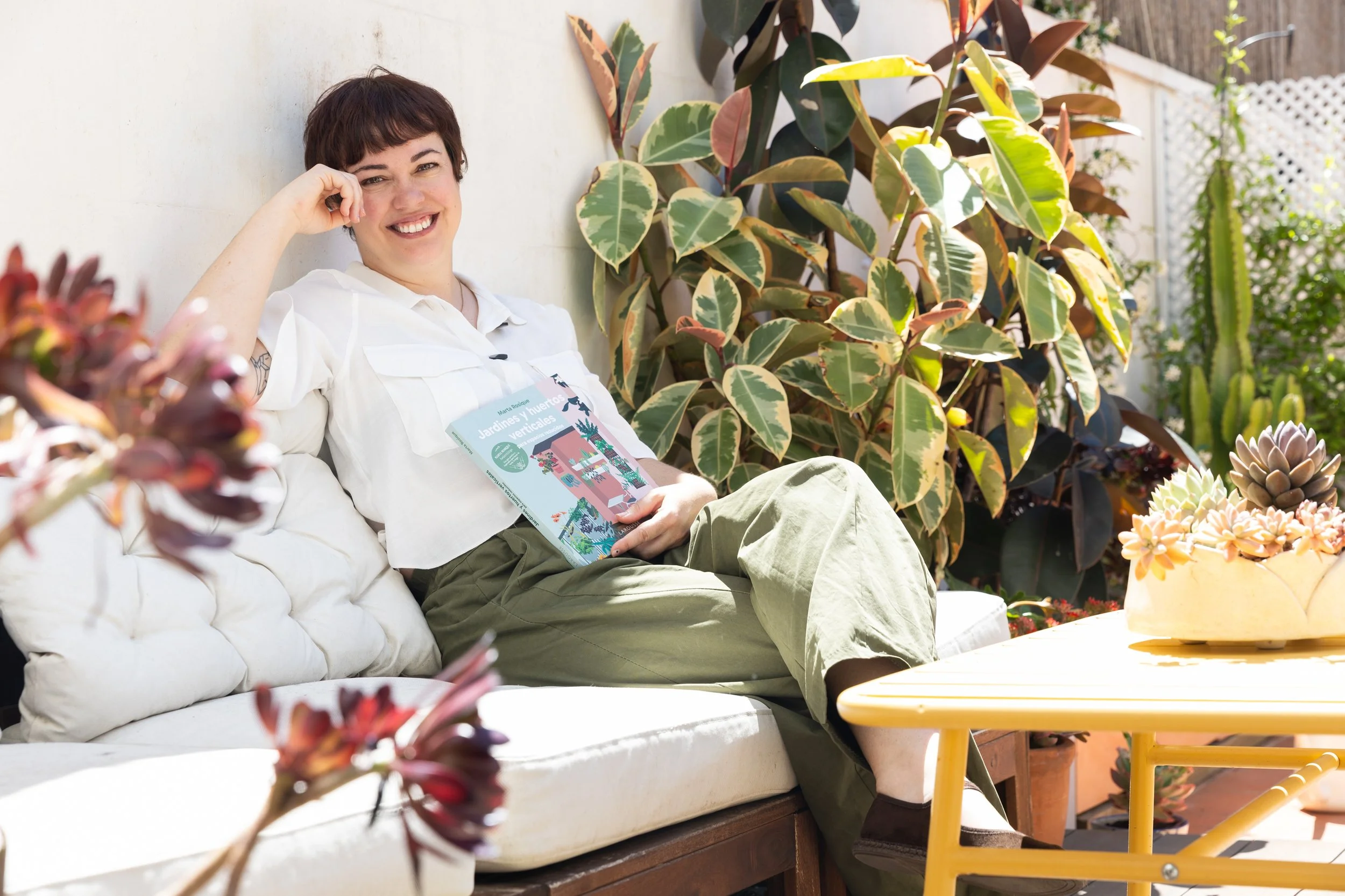 Mujer sonriendo mientras está sentada en un sofá en un patio con plantas verdes, sosteniendo un libro titulado 'Jardines y huertos verticales'.