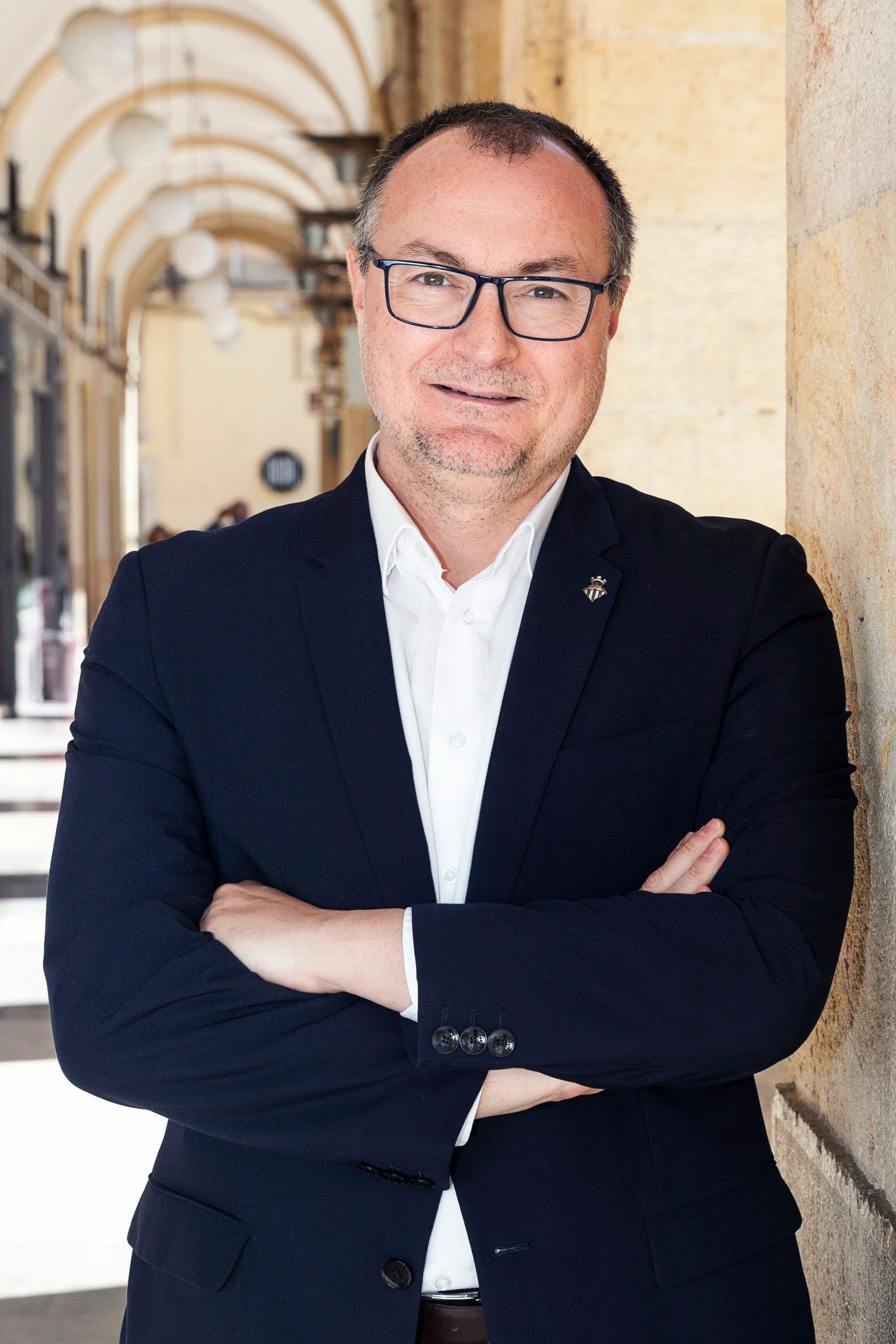 Hombre con gafas, ropa formal, de cabello corto y canoso, posando con los brazos cruzados en un pasillo con arcos y columnas de piedra clara.