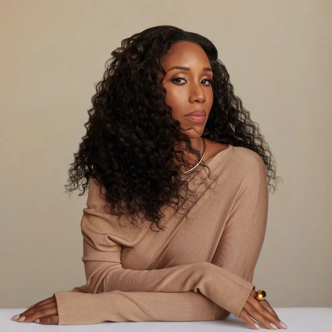 A woman with dark, curly hair and medium skin tone, wearing a beige long sleeve top, leaning on a white surface with her arm crossed, gazing confidently at the camera against a plain beige background.