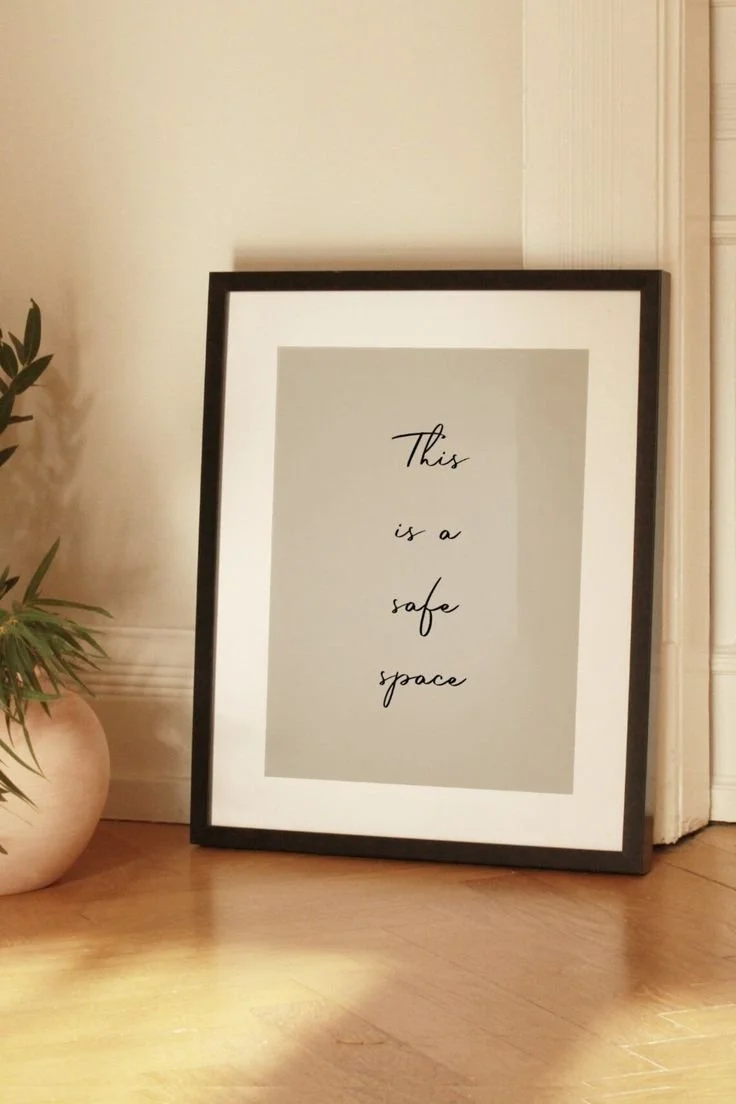 Framed sign on the floor that reads 'This is a safe space' leaning against a wall next to a potted plant.