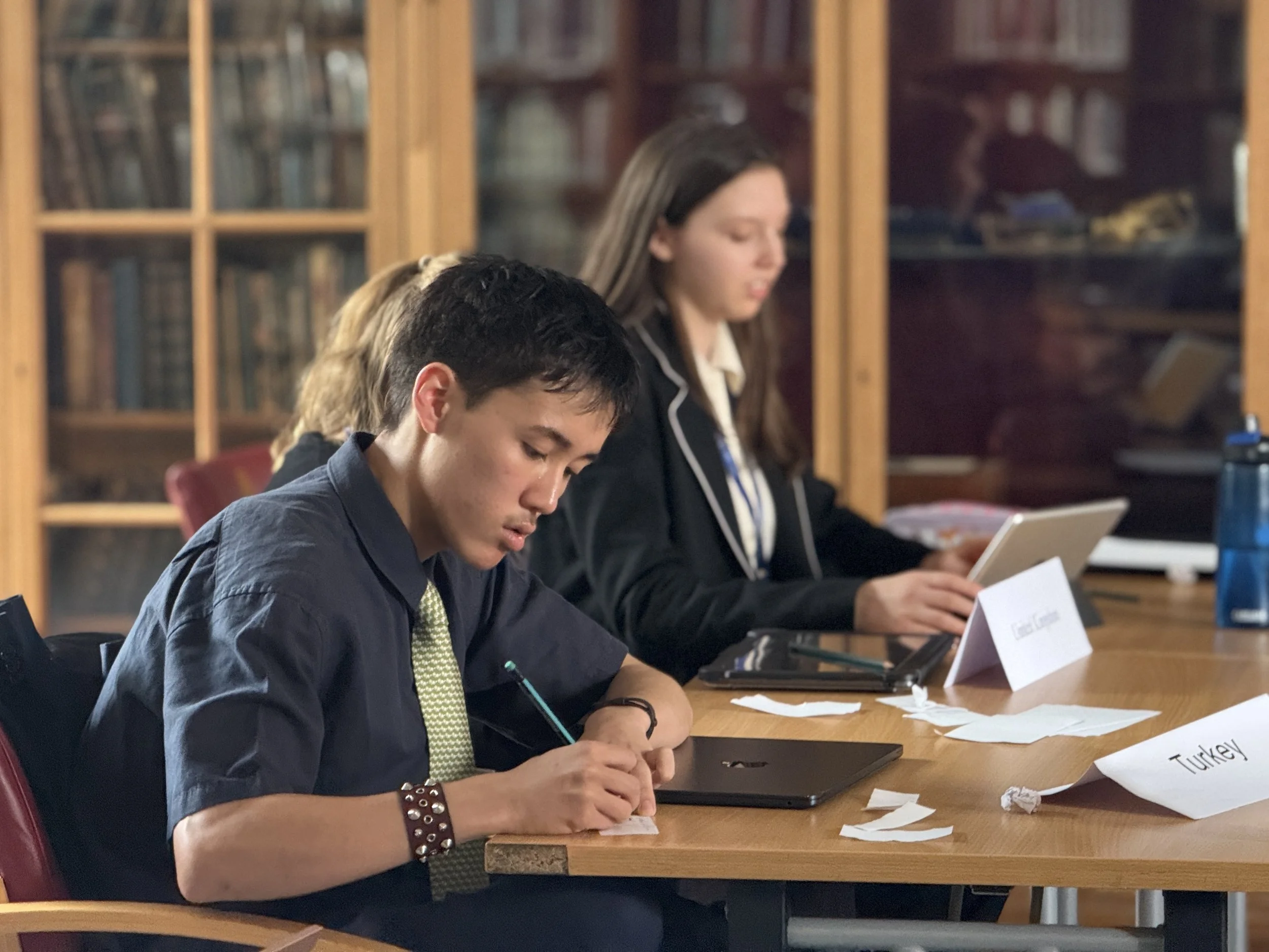 Young people in formal attire sitting at a conference table with nameplates and notes, engaged in a meeting in a library or conference room.