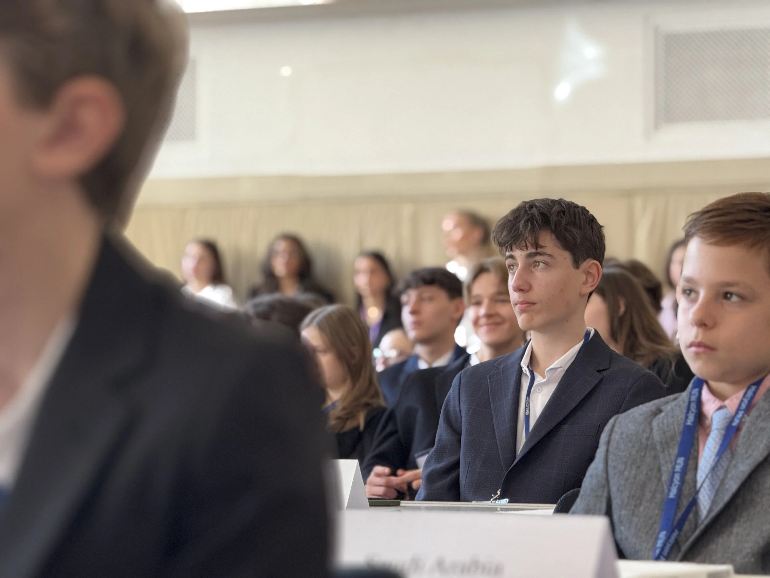 A group of students in formal attire attending a conference or seminar, seated in a room with beige walls and a high ceiling, listening attentively.