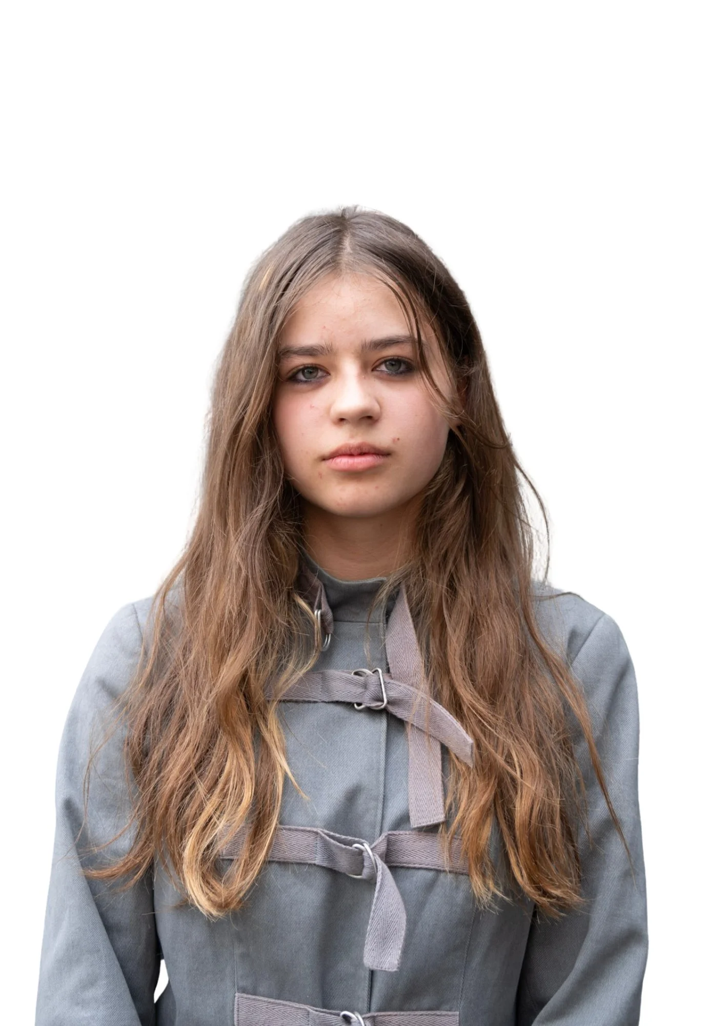A young woman with long, wavy brown hair and blue eyes wearing a gray coat with a tied belt, standing against a plain white background.