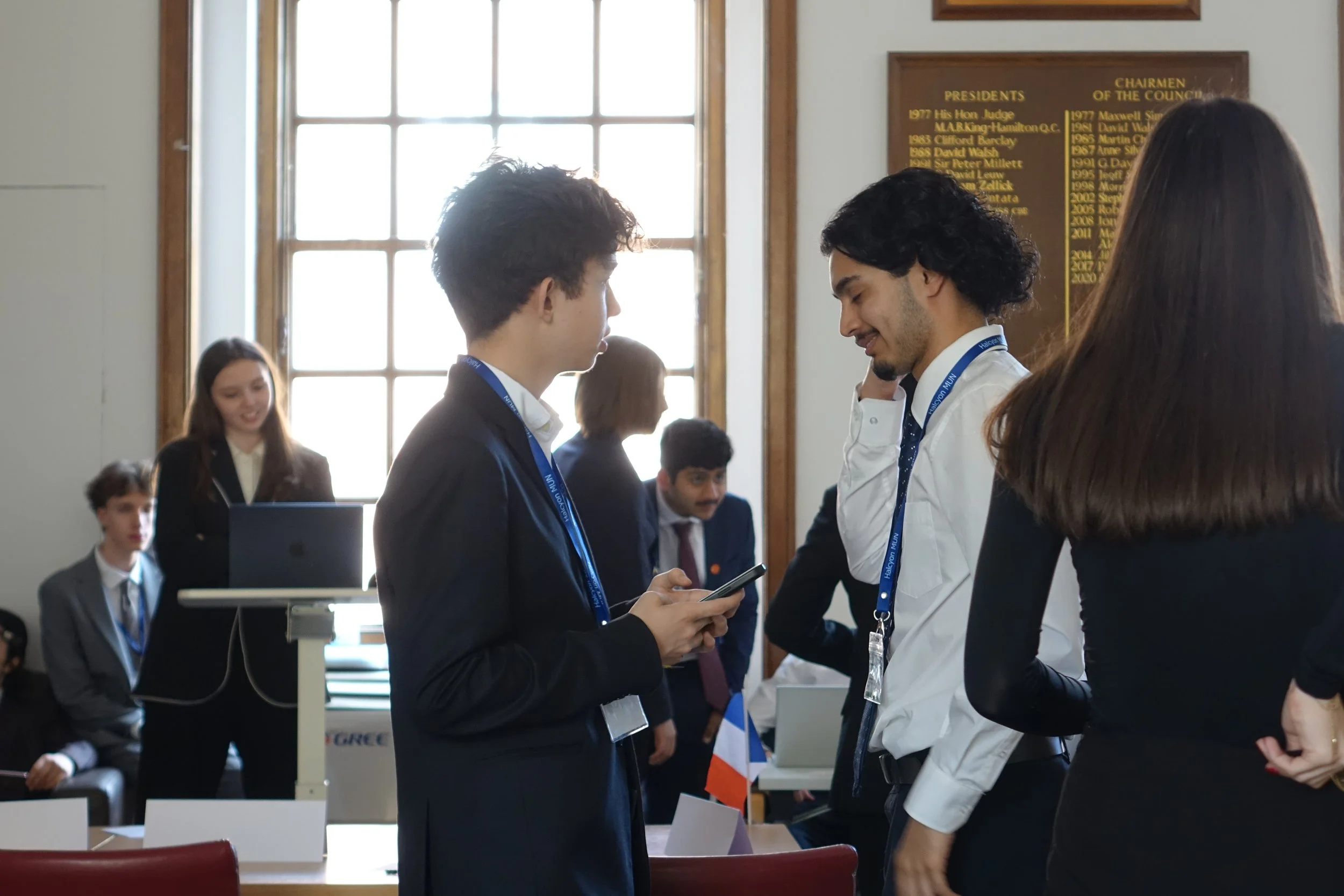 Young professionals conversing in a conference hall with large windows, a wooden plaque listing presidents and council chairs in the background, and a French flag on the table.