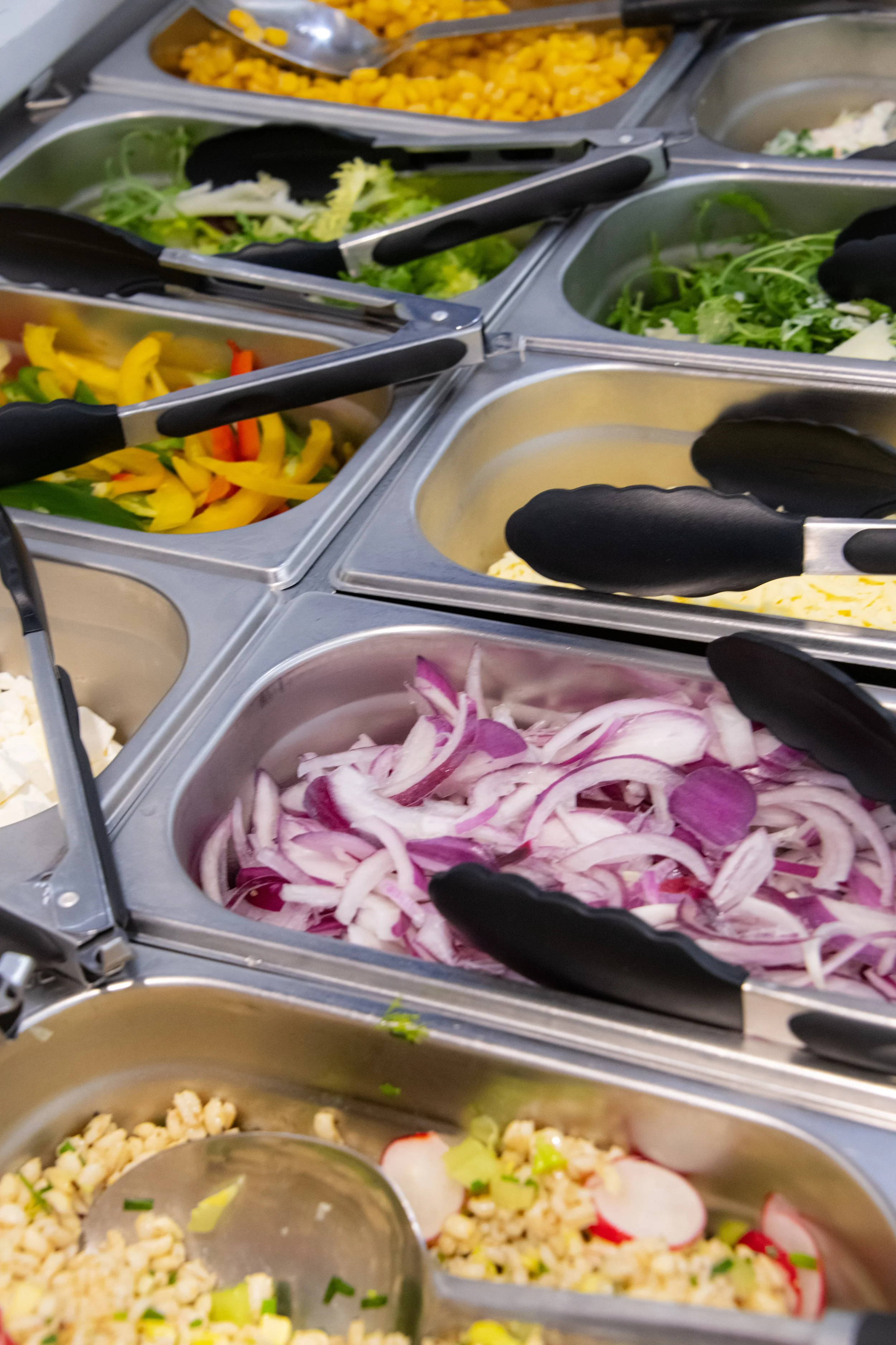 A buffet of fresh vegetables including sliced red onions, chopped radishes, chopped green onions, shredded cheese, bell peppers, lettuce, and corn in stainless steel serving trays with tongs.