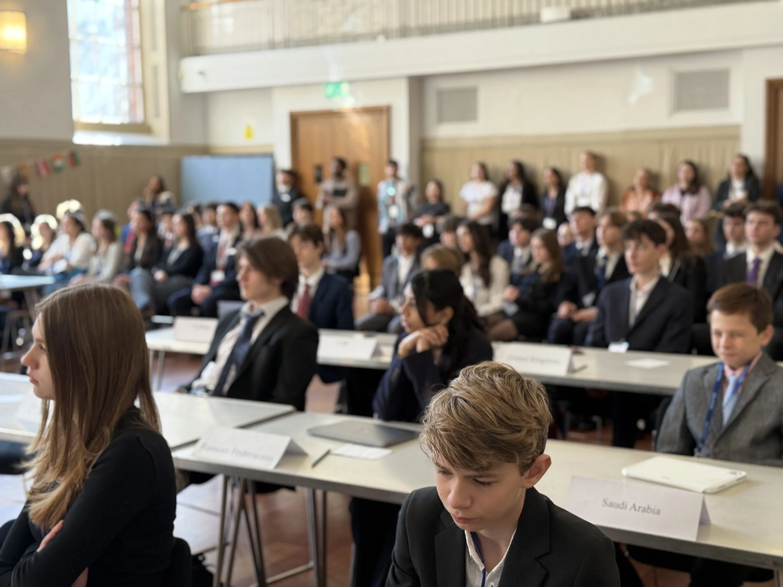 Students attending an international conference, seated at tables with country name cards, listening attentively, in a large well-lit room.