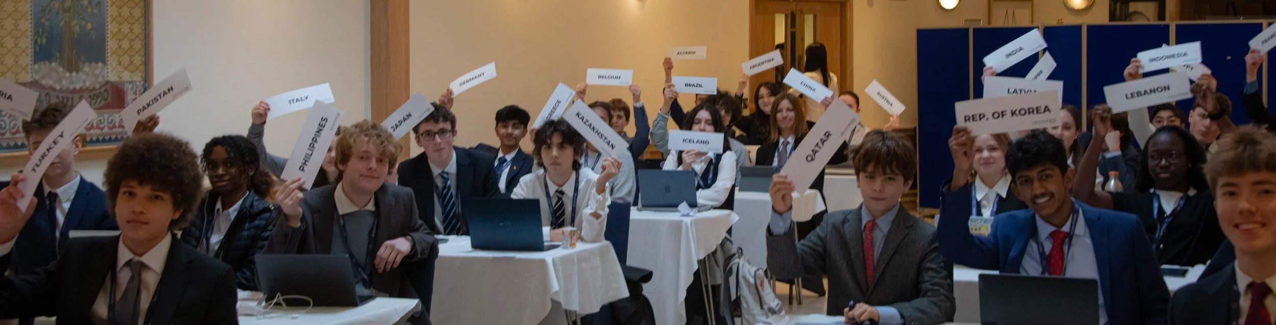 Students at a model United Nations conference holding signs with country names from various nations including Italy, Pakistan, Philippines, Turkey, Kazakhstan, Iceland, Qatar, Lebanon, South Korea, and others, seated at tables with laptops in a large room.