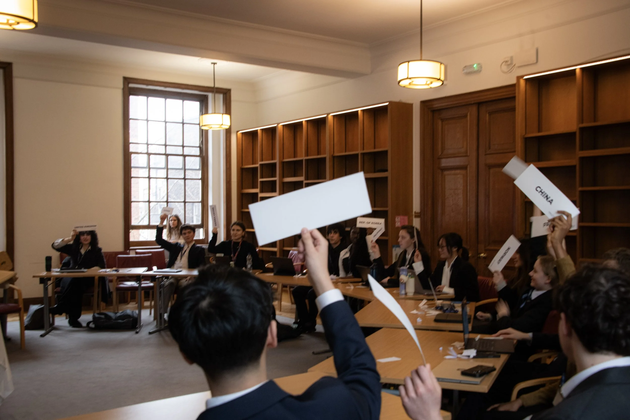 Students participating in a Model United Nations conference, raising paper signs with country names, in a formal conference room with wooden furniture and large windows.