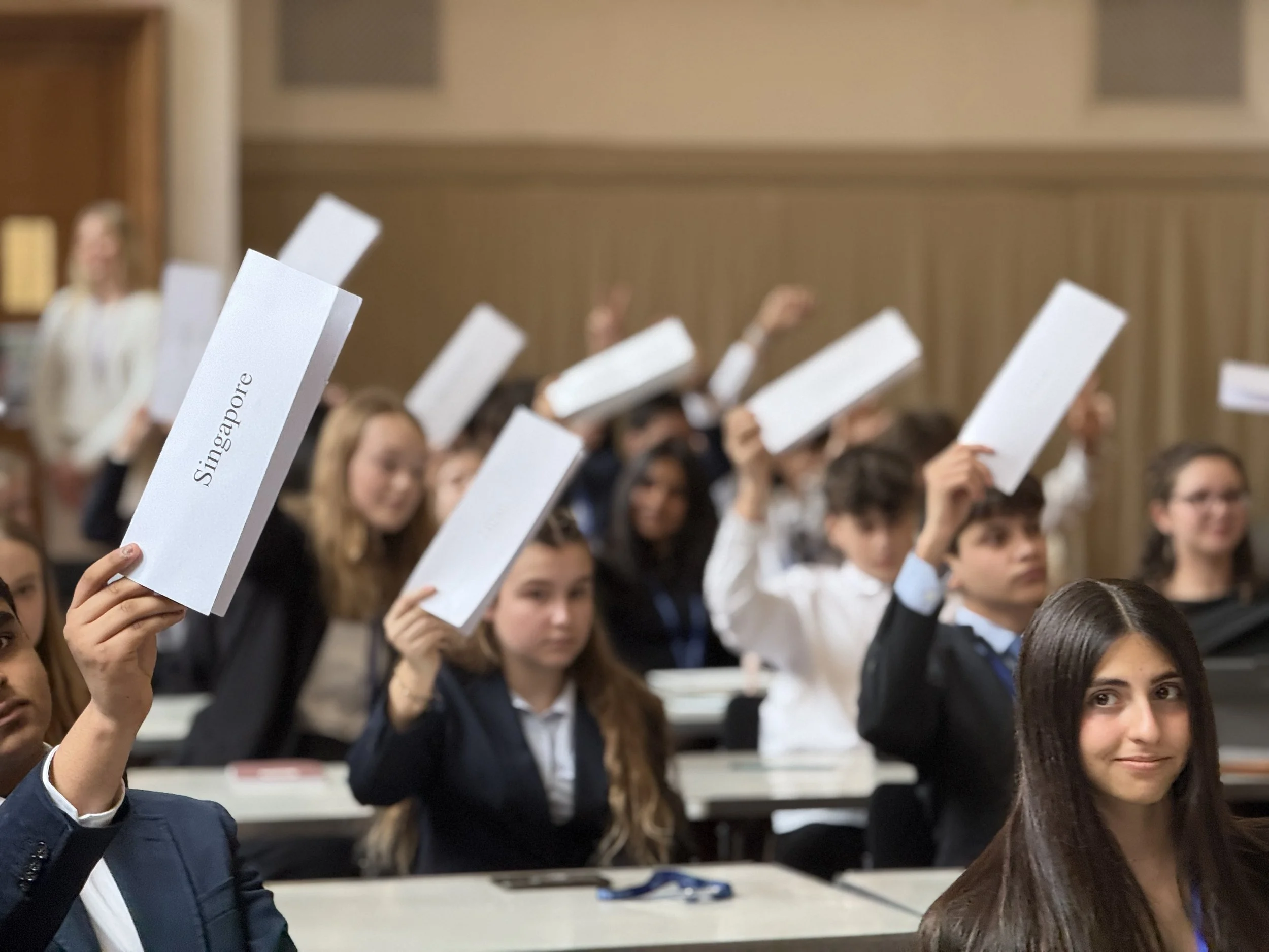 Students in a classroom with some holding up white papers labeled 'Singapore' and others raising their hands.
