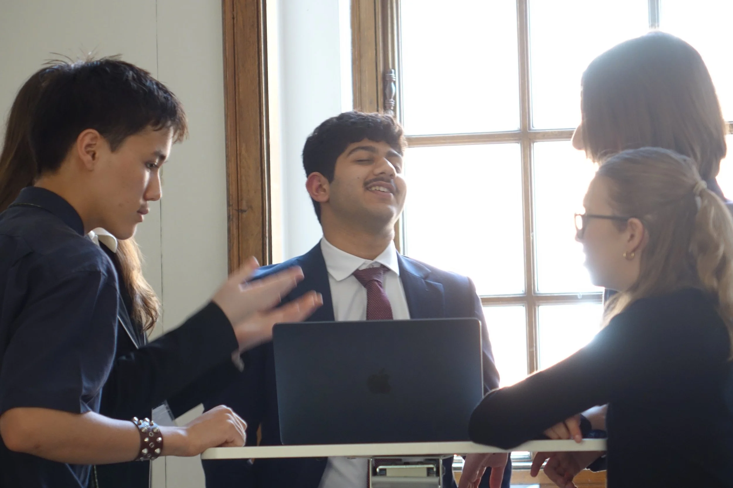 Group of four young professionals having a discussion around a table, with a laptop in front of them, near a large window.