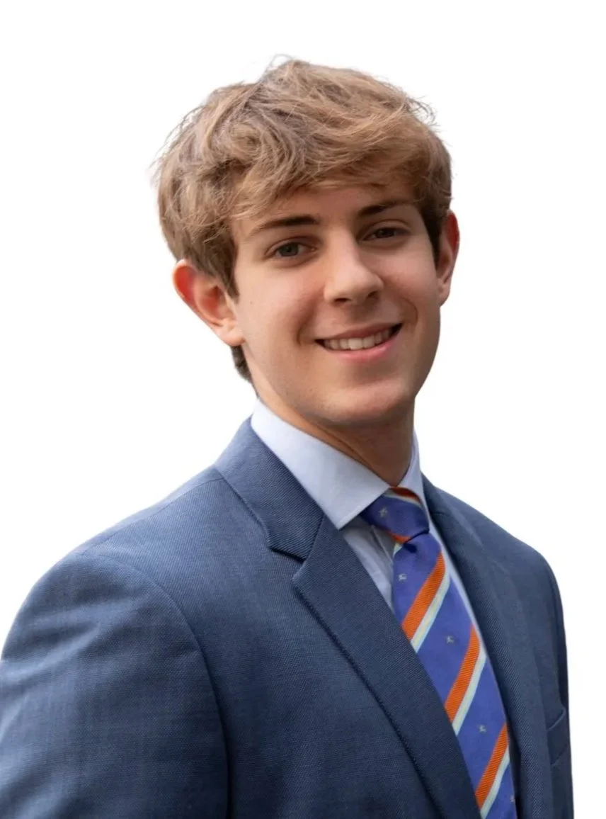 A young man with wavy, light brown hair smiling, dressed in a blue business suit with a light blue shirt and a colorful striped tie, standing against a plain white background.