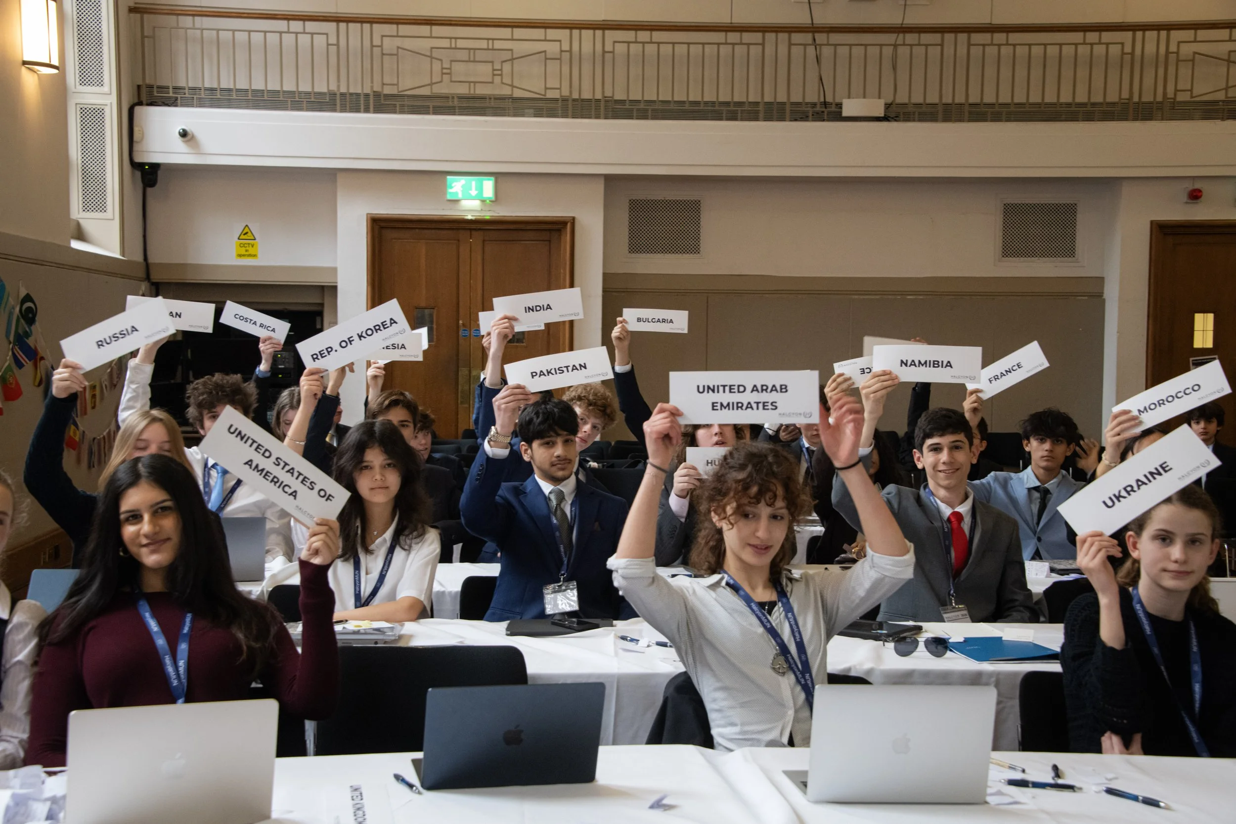 Young students seated at tables in a conference room holding up signs with country names, participating in an international mock assembly or quiz event.