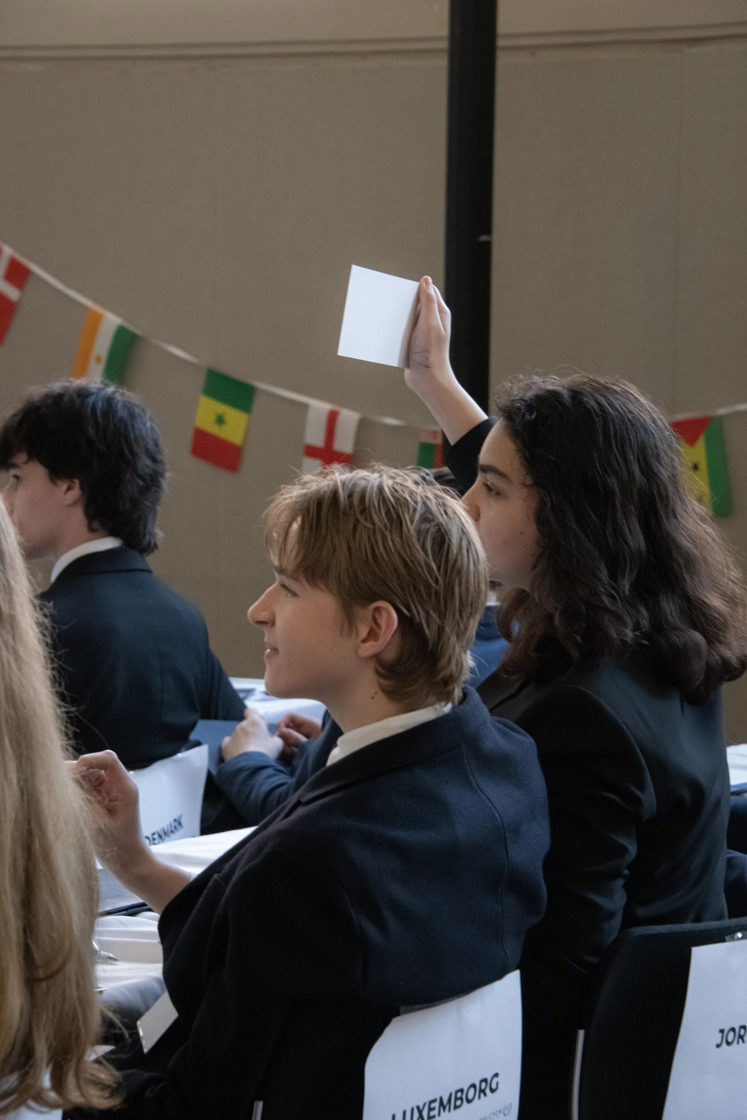 A girl is raising her hand during a conference or competition, with international flags hanging in the background. Other participants are seated in front of her, wearing formal attire.