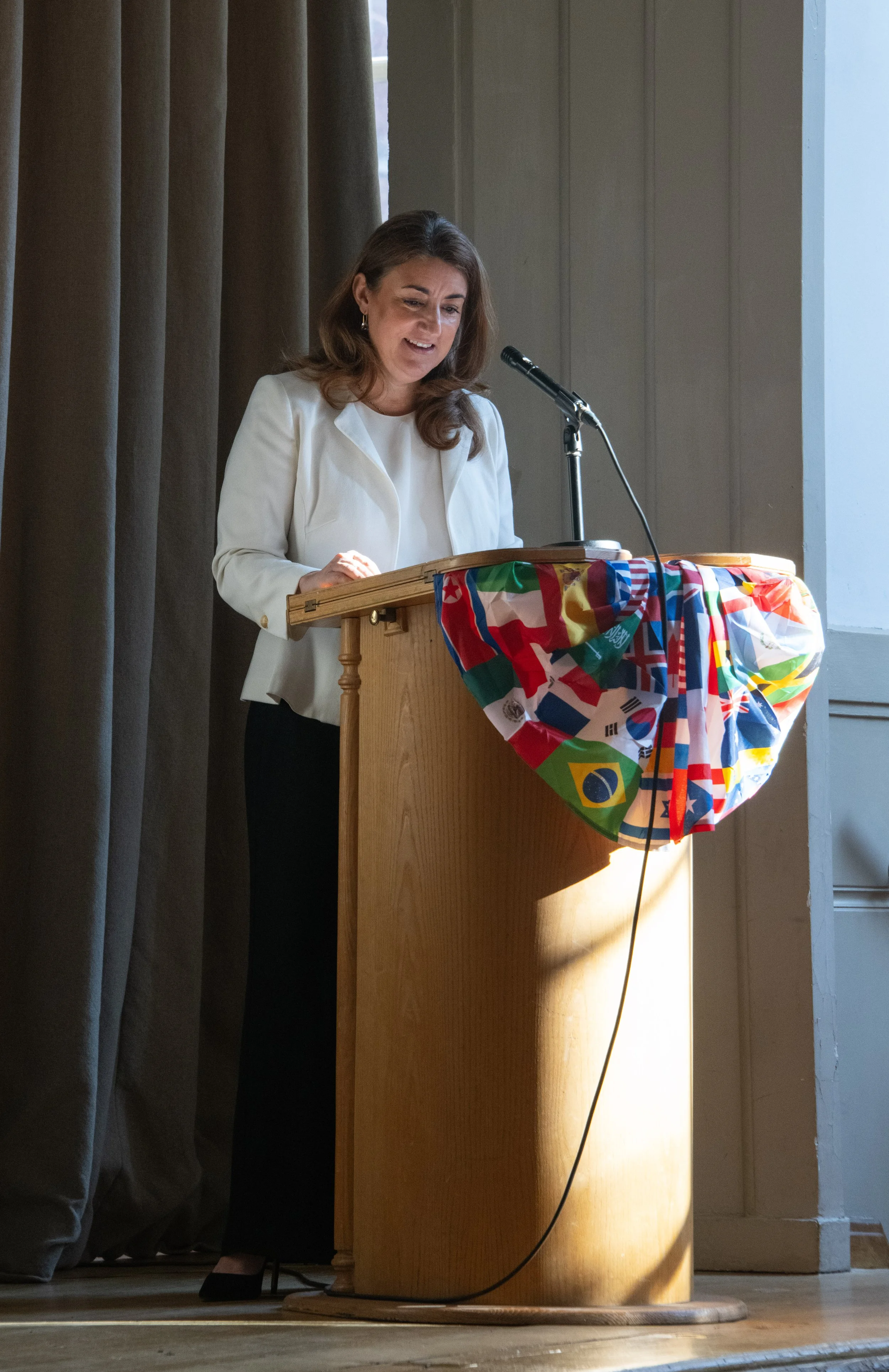 Woman in a white blazer speaking at a wooden podium draped with a colorful fabric with international flags, in a room with gray curtains and beige walls.