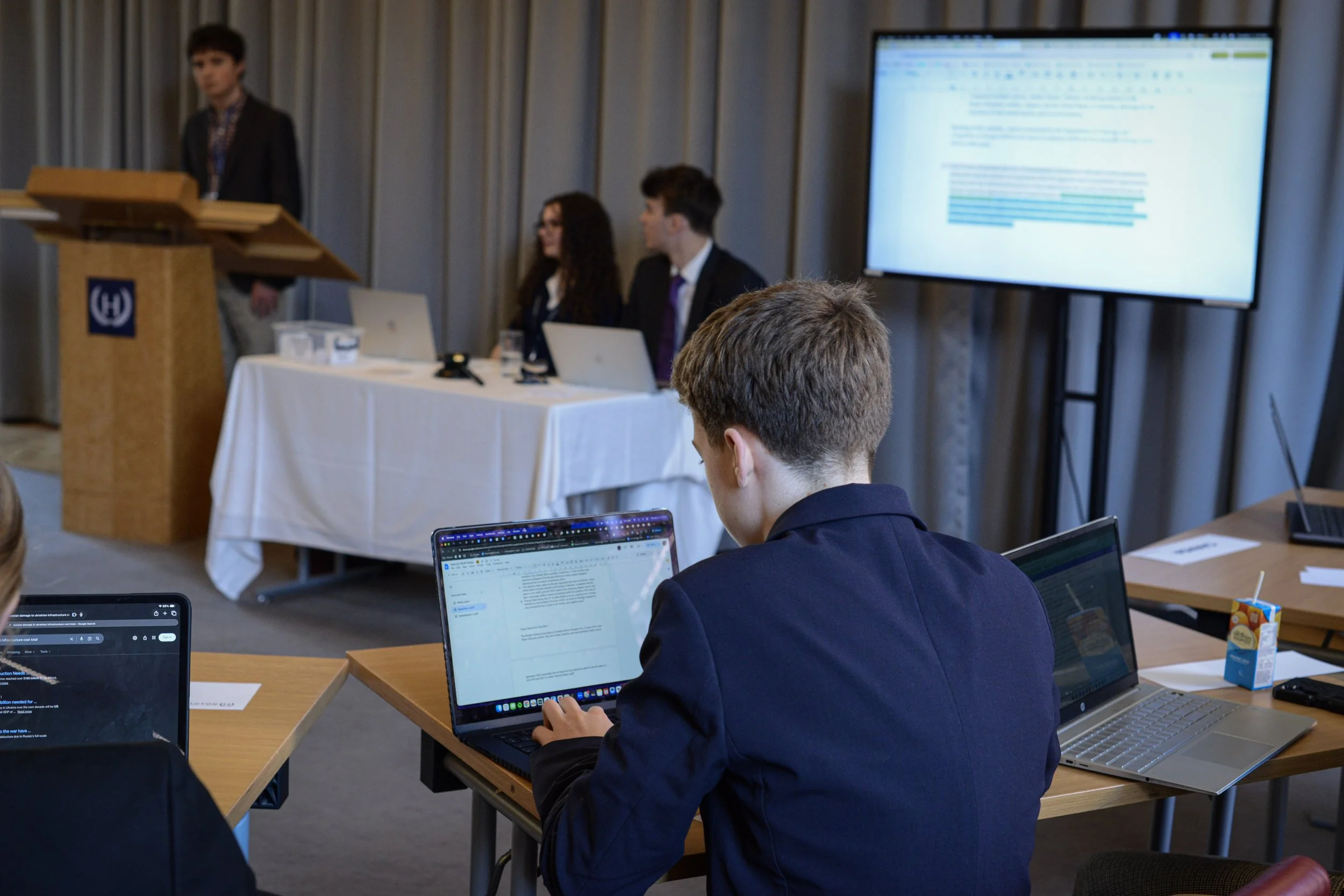A conference room with a speaker at a podium on the left, three people sitting at a table with laptops in the background, and a man in the foreground working on his laptop, with a large screen displaying a presentation on the right.